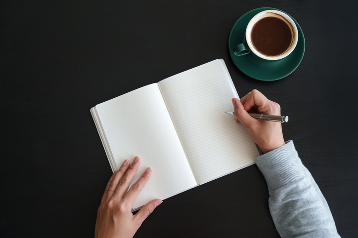 Overhead view of an Asian woman writing in diary while having coffee and working at the desk at home - stock photo